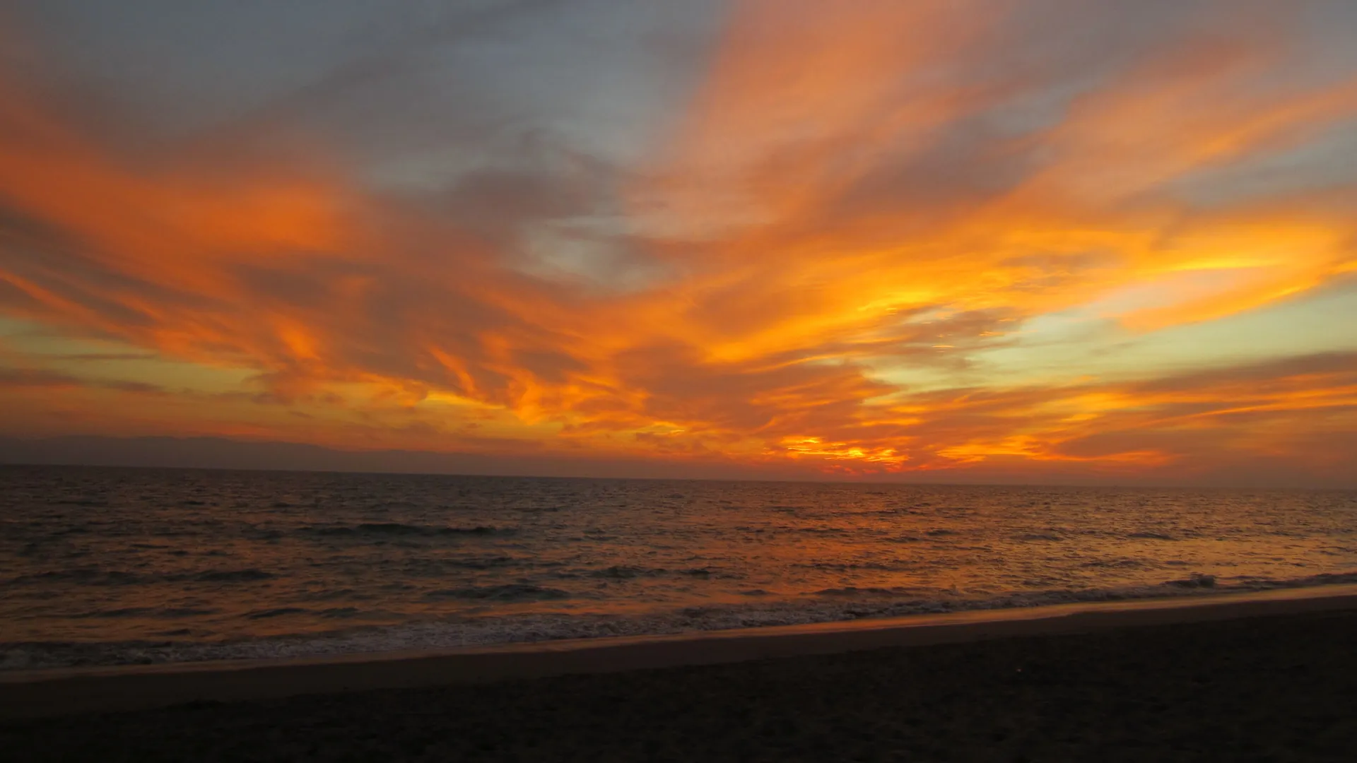Orange and gold streaks of cloud blaze across a sunset sky above a dark sea and a darkening beach on Banderas Bay