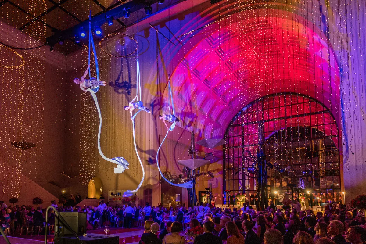 Aerialists hanging from white silks under a lit arched ceiling above a seated audience (an aerial silks act at a theatre in Texas, shown as an example of the art, not the Vallarta show)